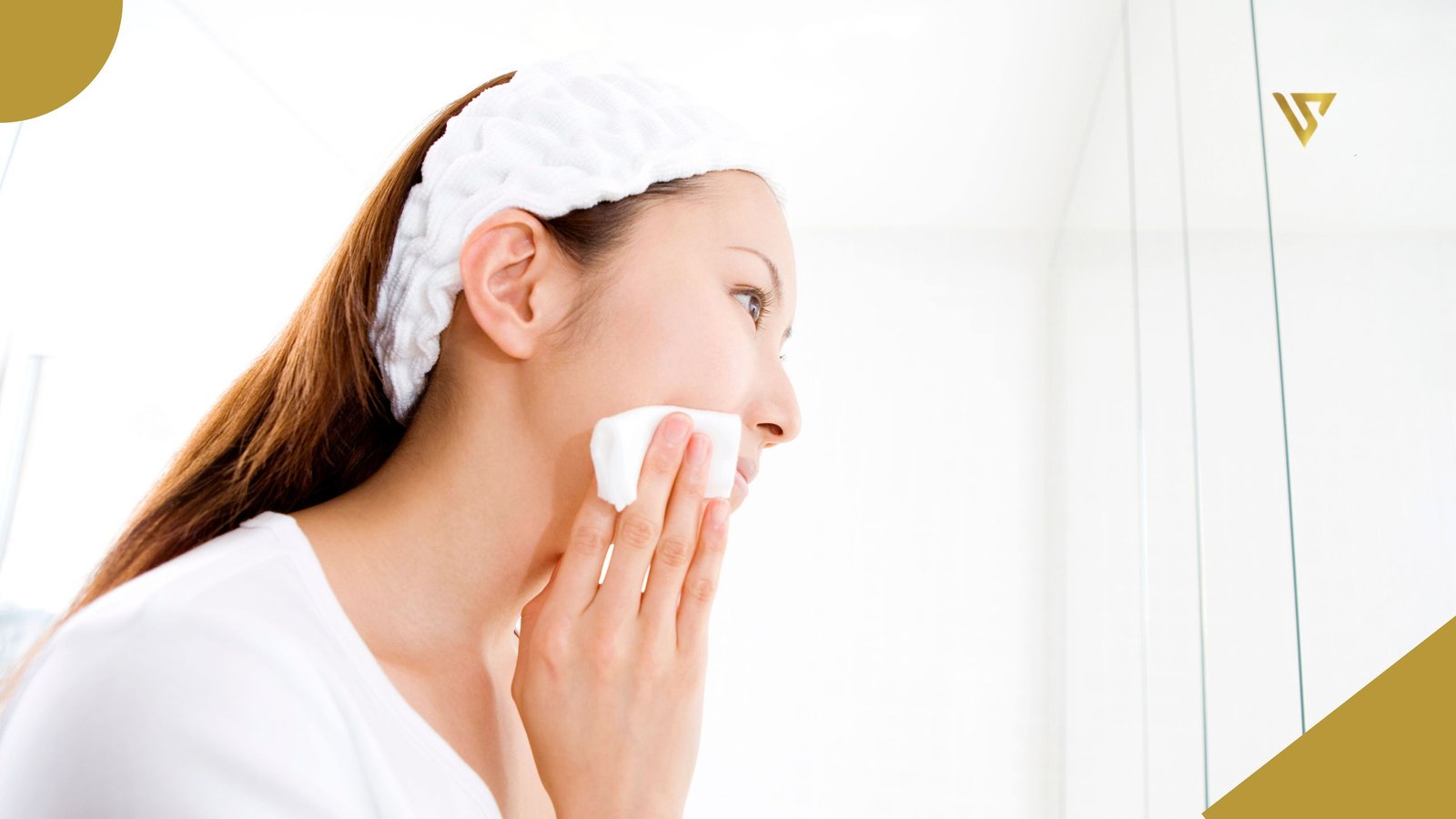 An adult woman with long brown hair, wearing a white shirt and a white headband, holding a makeup remover cloth in her right hand, resting it on her right cheek.