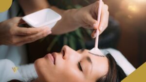 Right hand holding a white container, left arm hand holding a brush that is resting on the forehead of an adult woman with dark brown hair, who is lying on a treatment bed with her eyes closed, Chemical Peel for Glowing Skin: Your Safe Path to Holiday Radiance, beauty spa by val