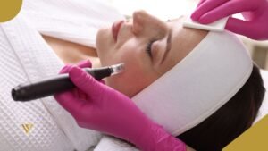 An adult woman with dark hair and closed eyes, lying down, wearing a white bathrobe and a white headband. A right hand wearing a fuchsia glove holds a small gauze pad resting on the woman’s forehead, while a left hand wearing a fuchsia glove holds a microneedling pen resting on the left cheek of the woman lying down Microneedling Facial Treatment Stimulate Collagen and Rejuvenate Your Skin Beauty spa by val
