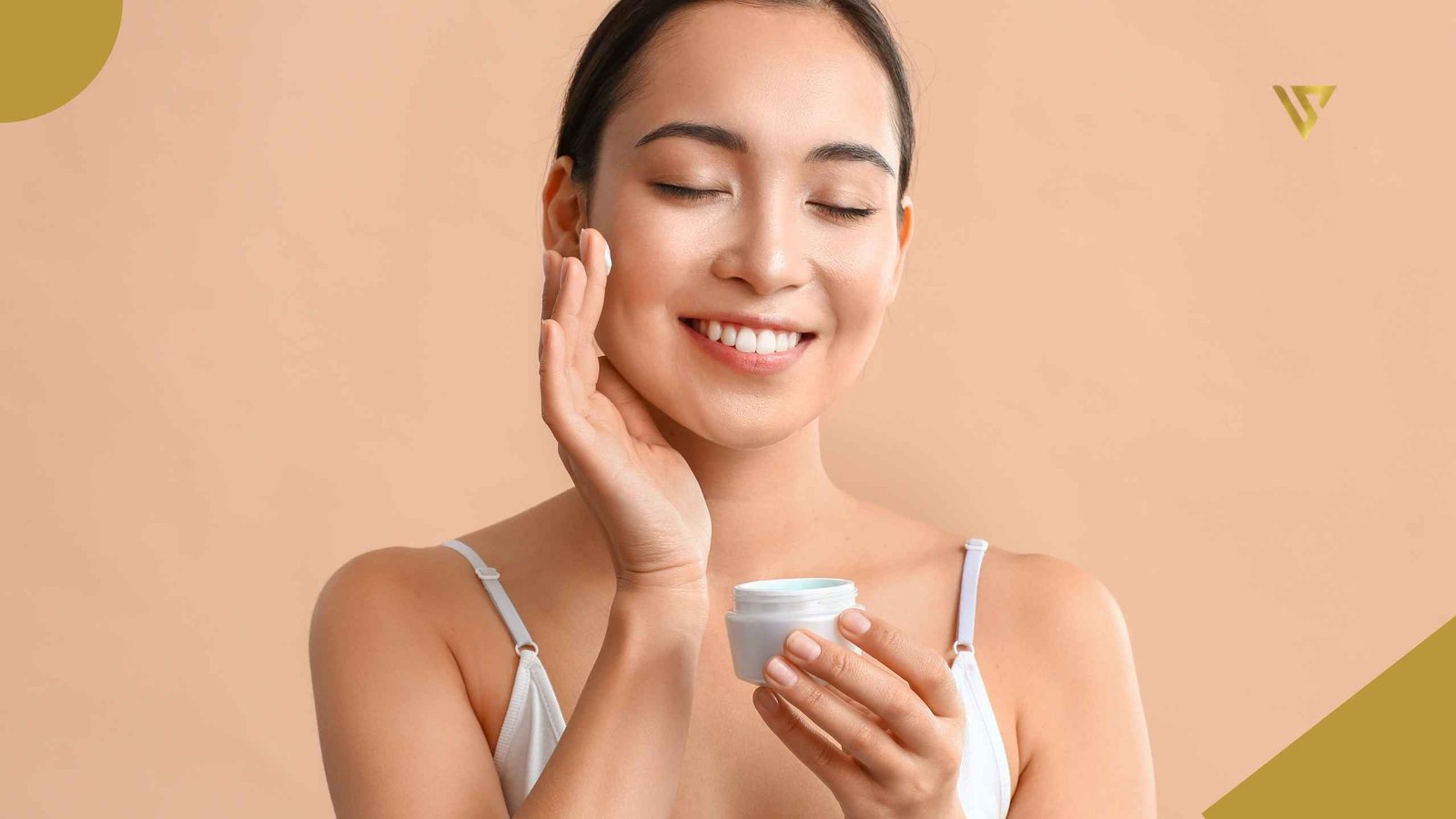 Woman with dark hair and closed eyes wearing a white strappy top. Her left hand holds a white container of cream, while her right hand is positioned near her right cheek. Her middle finger has cream on it and is resting against her right cheek. The background is light peach. How to Prepare Your Skin for Fall. Beauty Spa by Val