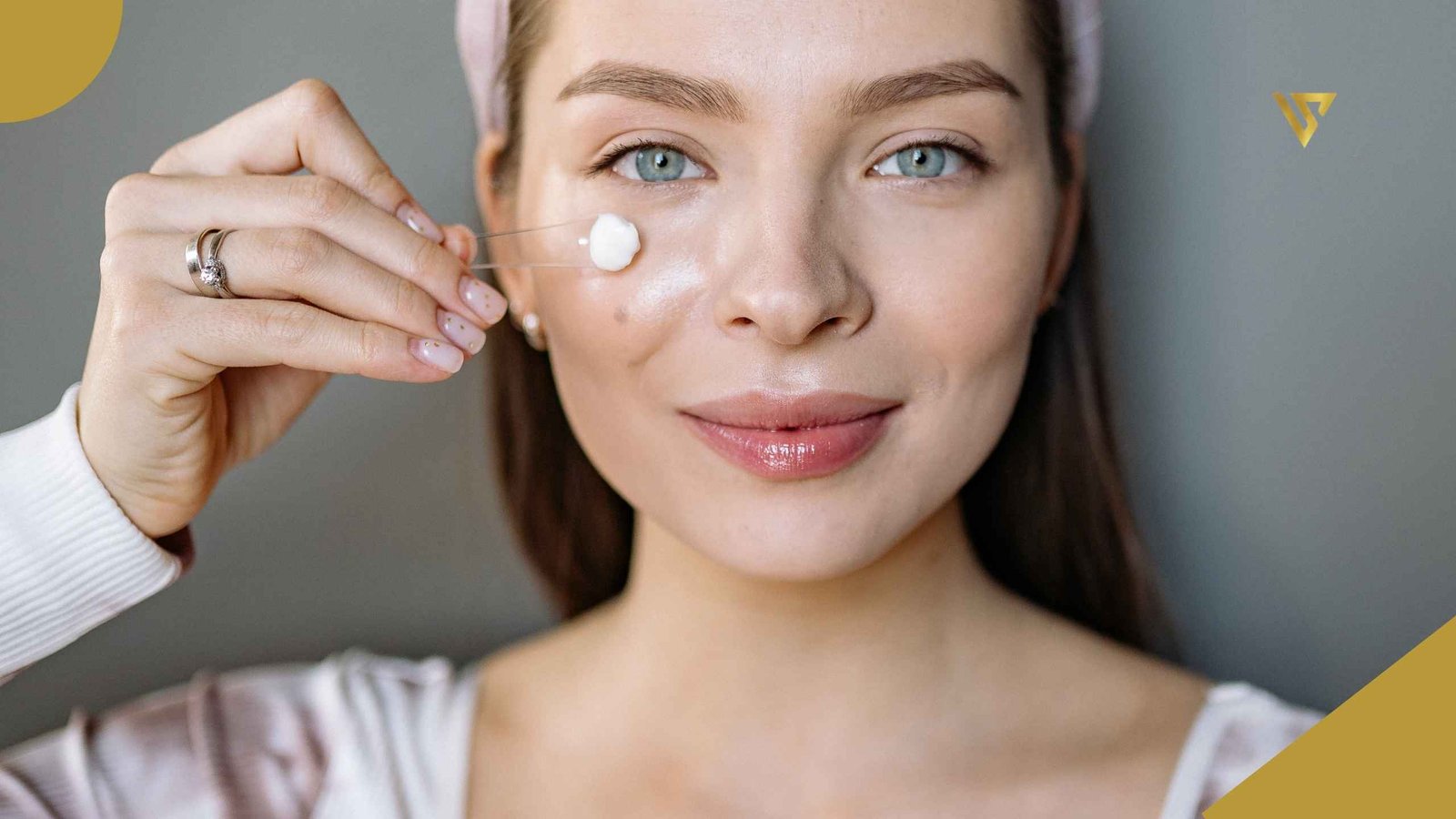 Woman with light-colored eyes and long hair wearing a headband and a white long-sleeved blouse. Her right hand has a silver wedding band and engagement ring on the ring finger. She holds a transparent spatula with white cream on the tip, resting against her cheek. Skin Barrier Repair: What It Is and How Spa Treatments Can Help, Beauty Spa by Val